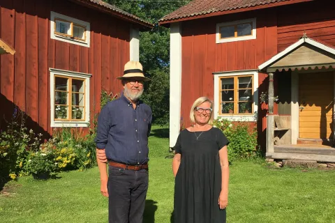 Äldre par poserar framför röda hus i traditionell stil i Dalarna. 